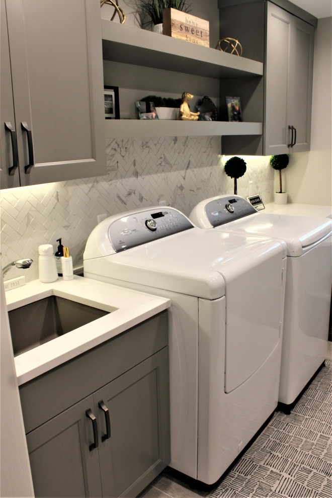 Inspiration for a large transitional u-shaped utility room in Detroit with an undermount sink, flat-panel cabinets, grey cabinets, quartz benchtops, grey splashback, stone tile splashback, grey walls, porcelain floors, a side-by-side washer and dryer, grey floor and white benchtop.