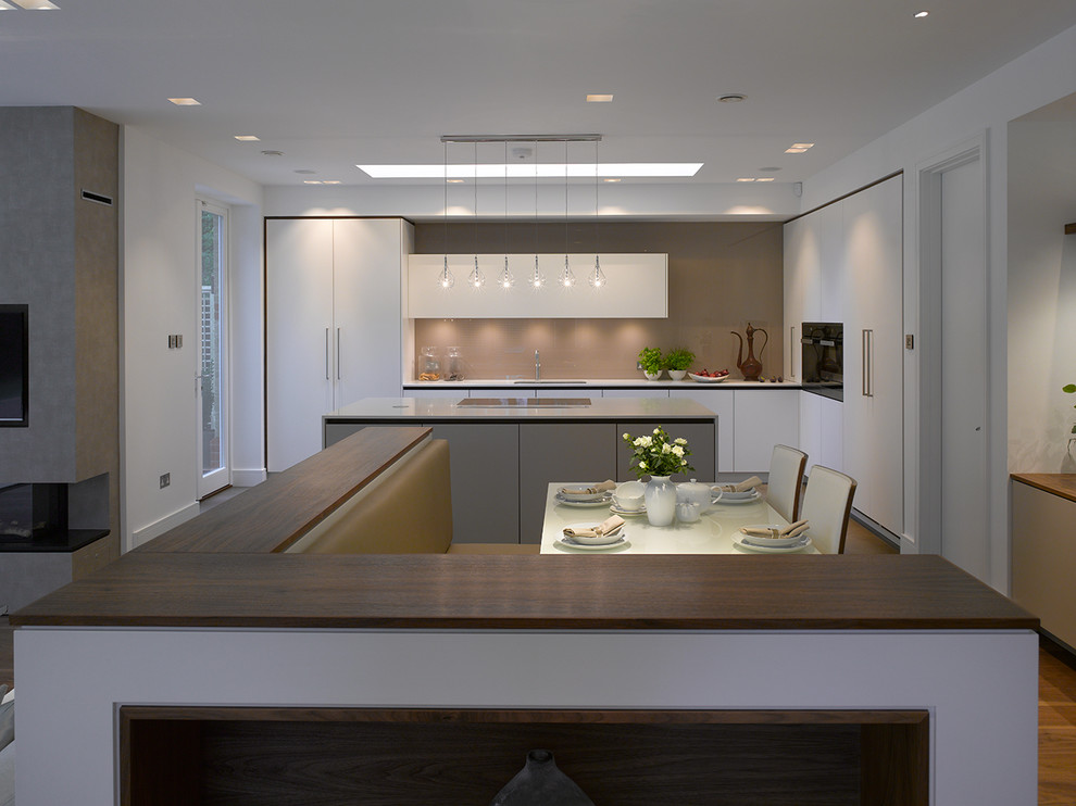 This is an example of an expansive contemporary open plan kitchen in London with flat-panel cabinets, white cabinets, solid surface benchtops, beige splashback, glass sheet splashback, with island and white benchtop.