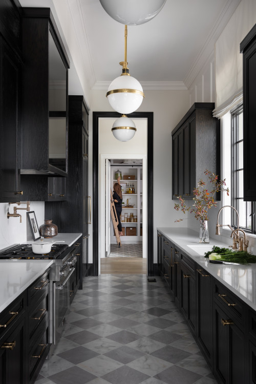 Galley kitchen with parallel black cabinets, white countertops, brass hardware, globe pendants, and a central corridor leading to a walk-in pantry