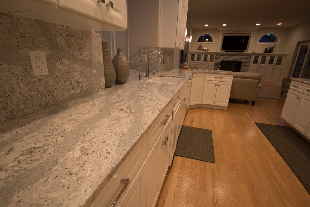 Large transitional l-shaped medium tone wood floor open concept kitchen photo in Seattle with an undermount sink, recessed-panel cabinets, white cabinets, quartz countertops, gray backsplash, stone slab backsplash, white appliances and an island