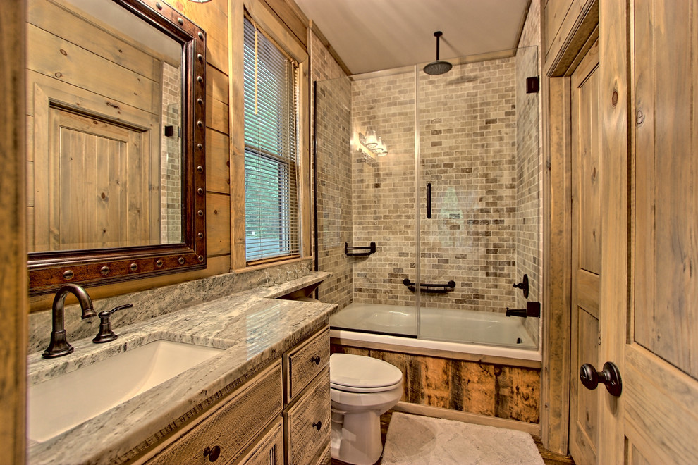 Photo of a small country master bathroom in Other with recessed-panel cabinets, distressed cabinets, a drop-in tub, a shower/bathtub combo, a two-piece toilet, multi-coloured tile, stone tile, grey walls, painted wood floors, a drop-in sink, granite benchtops, brown floor and a hinged shower door.