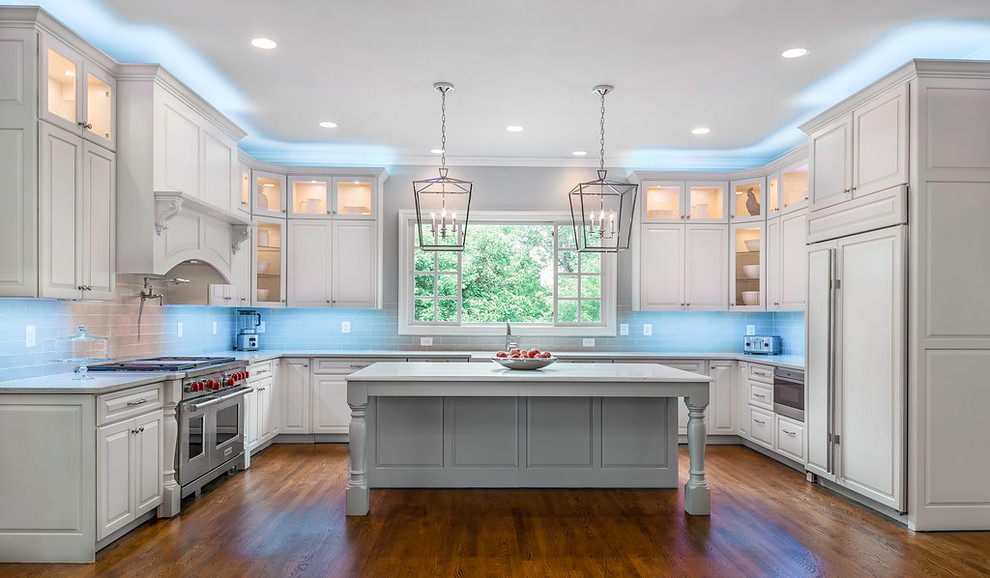 This is an example of an expansive traditional u-shaped eat-in kitchen in DC Metro with a farmhouse sink, raised-panel cabinets, white cabinets, quartz benchtops, grey splashback, glass tile splashback, stainless steel appliances, medium hardwood floors, with island, brown floor and white benchtop.