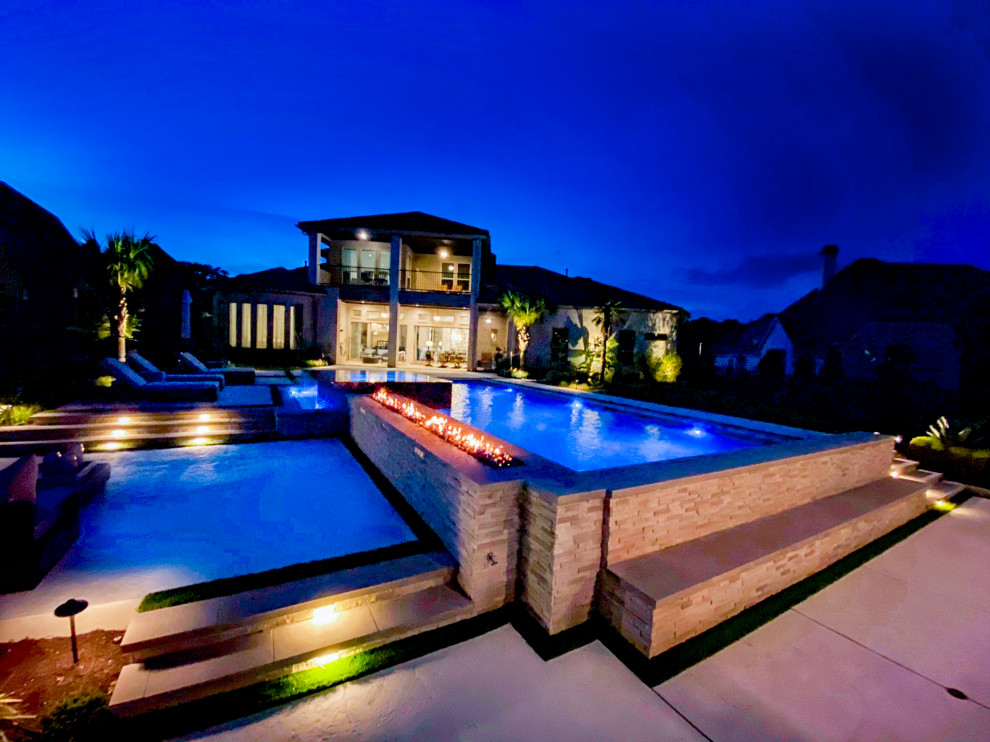 This is an example of a large modern backyard custom-shaped infinity pool in Dallas with a water feature and stamped concrete.