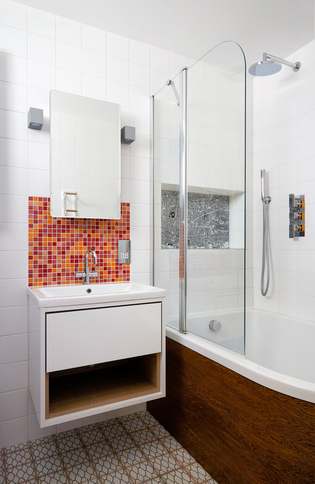 This is an example of a small contemporary kids bathroom in London with flat-panel cabinets, white cabinets, a drop-in tub, a shower/bathtub combo, a one-piece toilet, white tile, grey walls, ceramic floors, an integrated sink and multi-coloured floor.
