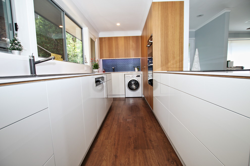 Photo of a mid-sized modern u-shaped kitchen pantry in Sydney with a double-bowl sink, flat-panel cabinets, white cabinets, quartz benchtops, white splashback, stone slab splashback, stainless steel appliances, vinyl floors, a peninsula and brown floor.