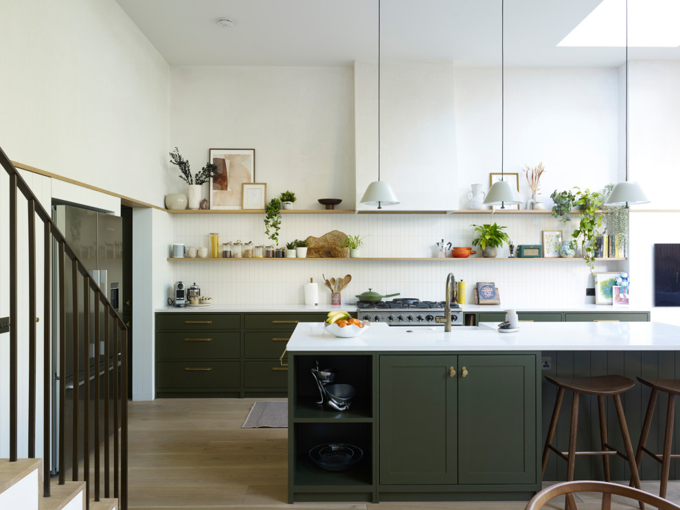 Photo of a transitional galley eat-in kitchen in London with a farmhouse sink, flat-panel cabinets, green cabinets, solid surface benchtops, white splashback, cement tile splashback, stainless steel appliances, medium hardwood floors, with island, beige floor and white benchtop.