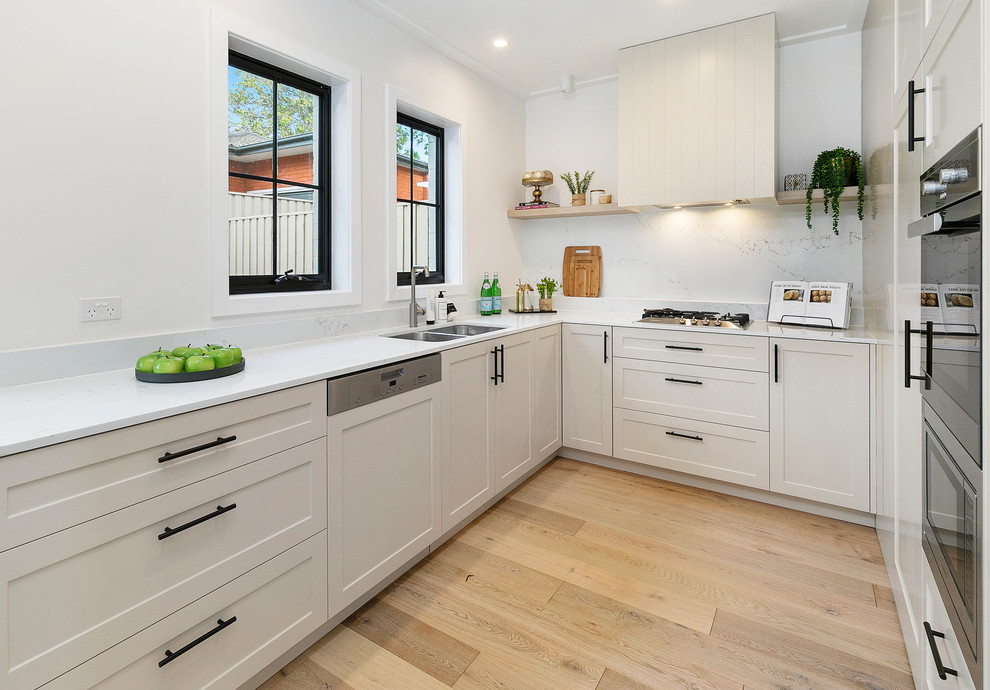 This is an example of a small transitional l-shaped open plan kitchen in Sydney with a double-bowl sink, white cabinets, marble benchtops, marble splashback, stainless steel appliances, light hardwood floors, white benchtop, recessed-panel cabinets, white splashback, multiple islands and brown floor.