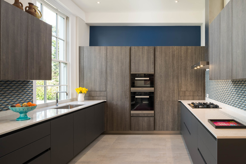 Inspiration for a large contemporary u-shaped open plan kitchen in London with a single-bowl sink, flat-panel cabinets, grey cabinets, solid surface benchtops, blue splashback, porcelain splashback, black appliances, ceramic floors, no island and grey floor.