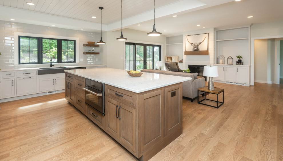 Inspiration for a large transitional l-shaped open plan kitchen in Milwaukee with a farmhouse sink, shaker cabinets, medium wood cabinets, quartz benchtops, white splashback, porcelain splashback, black appliances, light hardwood floors, with island, brown floor, white benchtop and timber.
