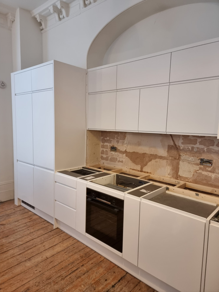 Large modern single-wall open plan kitchen in London with a built-in sink, flat-panel cabinets, white cabinets, quartz worktops, white splashback, stone slab splashback, white appliances, painted wood flooring and white worktops.