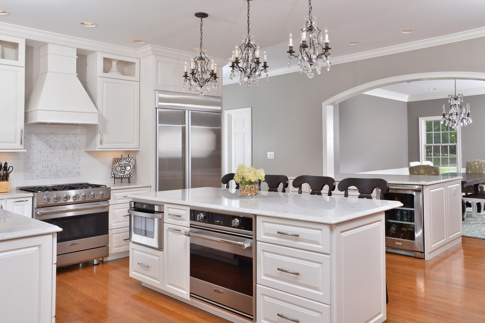 This is an example of a mid-sized traditional l-shaped eat-in kitchen in Cleveland with raised-panel cabinets, white cabinets, granite benchtops, white splashback, stainless steel appliances, medium hardwood floors, with island and brown floor.