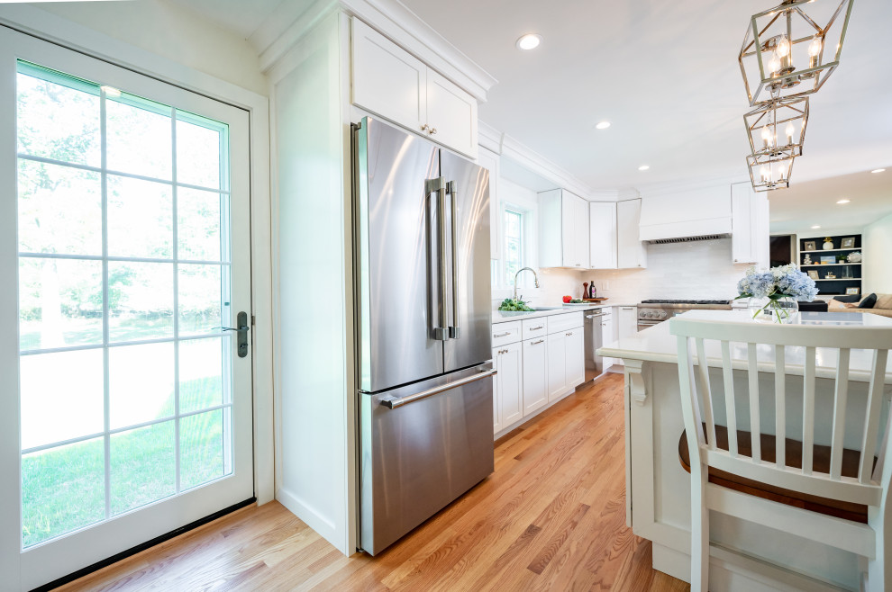 This is an example of a large transitional l-shaped kitchen pantry in Baltimore with an undermount sink, shaker cabinets, white cabinets, quartz benchtops, white splashback, ceramic splashback, stainless steel appliances, light hardwood floors, with island, brown floor and white benchtop.