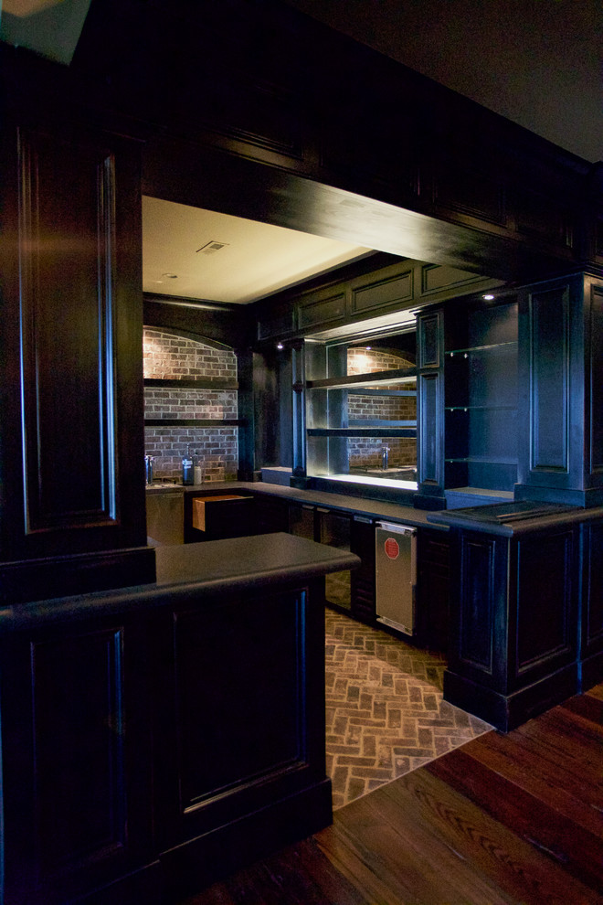 This is an example of a traditional galley seated home bar in Other with raised-panel cabinets, black cabinets, granite benchtops, brown splashback, stone tile splashback and brick floors.