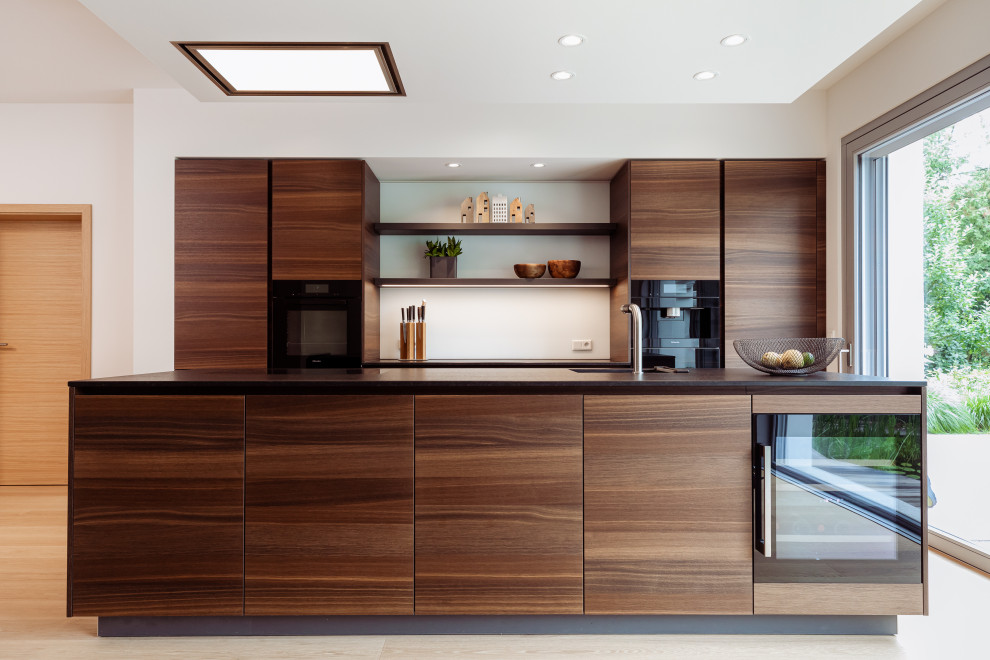 Design ideas for a mid-sized contemporary galley open plan kitchen in Frankfurt with an undermount sink, flat-panel cabinets, dark wood cabinets, granite benchtops, white splashback, glass sheet splashback, black appliances, light hardwood floors, with island and black benchtop.