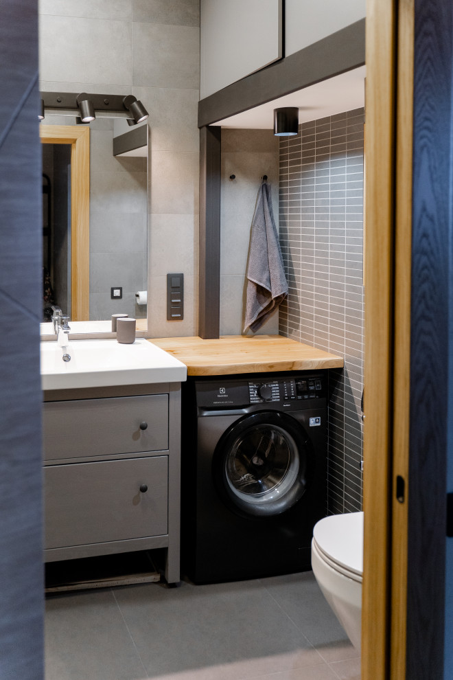 Small industrial bathroom in Saint Petersburg with flat-panel cabinets, grey cabinets, a wall-mount toilet, gray tile, porcelain tile, ceramic floors, wood benchtops, grey floor, a laundry, a single vanity and a freestanding vanity.