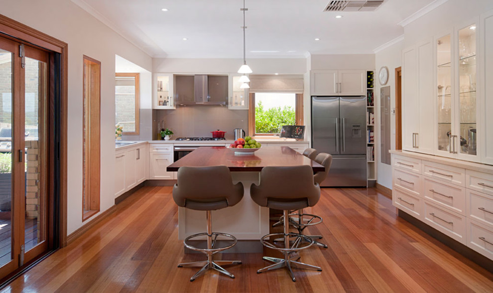 Photo of a mid-sized traditional l-shaped open plan kitchen in Melbourne with a double-bowl sink, shaker cabinets, white cabinets, quartz benchtops, beige splashback, glass sheet splashback, stainless steel appliances, light hardwood floors and with island.