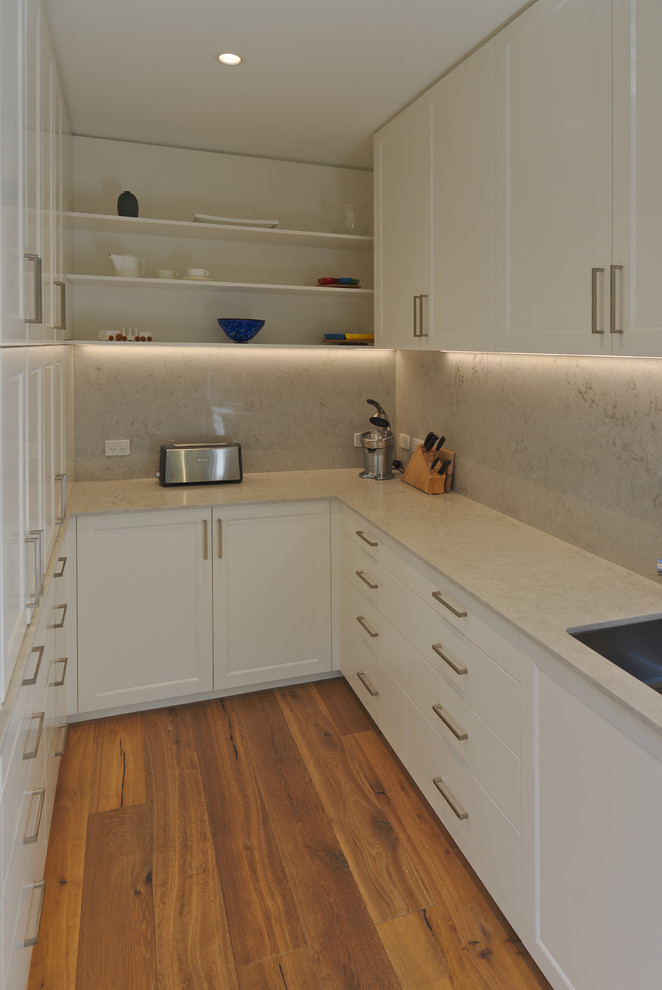 Inspiration for a mid-sized transitional galley open plan kitchen in Melbourne with an undermount sink, recessed-panel cabinets, white cabinets, quartz benchtops, beige splashback, stone slab splashback, black appliances, medium hardwood floors, with island, brown floor and beige benchtop.