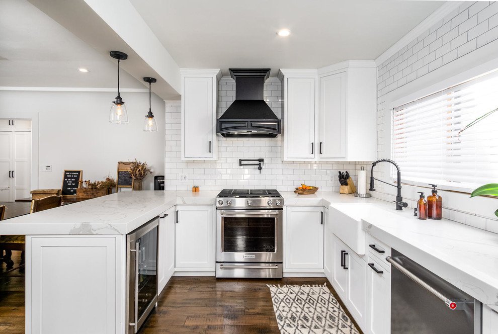 Design ideas for a traditional u-shaped kitchen in San Diego with a farmhouse sink, shaker cabinets, white cabinets, white splashback, subway tile splashback, stainless steel appliances, dark hardwood floors, a peninsula, brown floor and white benchtop.