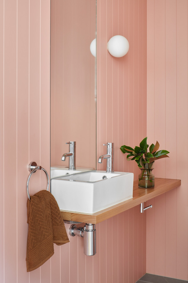 На фото: ванная комната среднего размера в современном стиле с фасадами цвета дерева среднего тона, розовыми стенами, раковиной с пьедесталом, столешницей из дерева, коричневой столешницей, тумбой под одну раковину, подвесной тумбой и панелями на части стены с