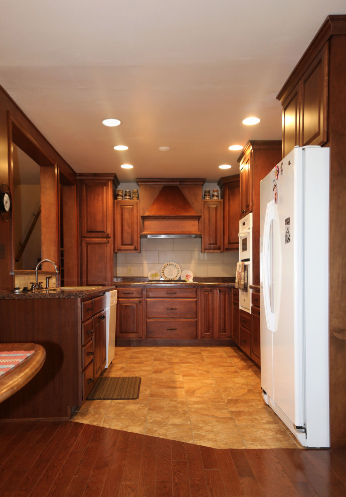 Photo of a mid-sized traditional u-shaped eat-in kitchen in Denver with an undermount sink, raised-panel cabinets, brown cabinets, granite benchtops, white splashback, ceramic splashback, white appliances, ceramic floors, with island, beige floor and brown benchtop.