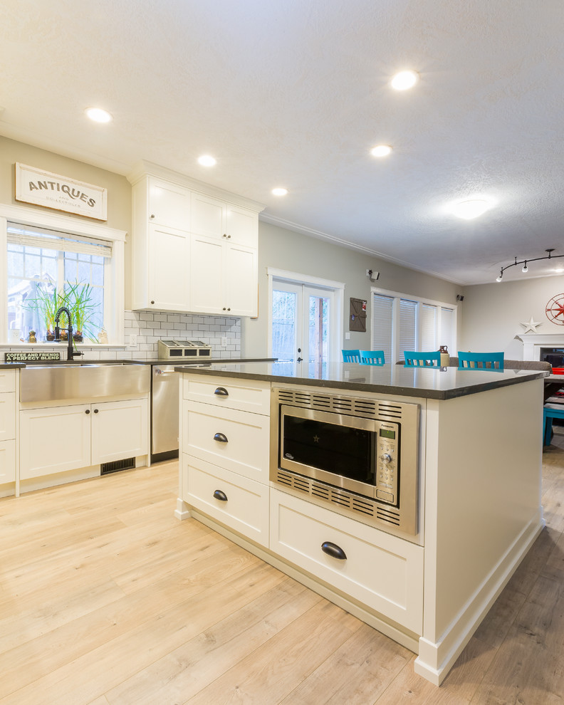 Photo of a large country l-shaped open plan kitchen in Other with a farmhouse sink, shaker cabinets, white cabinets, quartz benchtops, white splashback, subway tile splashback, stainless steel appliances, light hardwood floors, with island and black benchtop.