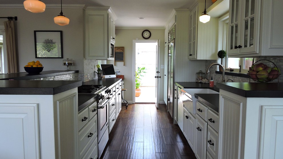 Photo of a beach style galley eat-in kitchen in Santa Barbara with no island, recessed-panel cabinets, white cabinets, granite benchtops, grey splashback, mosaic tile splashback, stainless steel appliances, a farmhouse sink and medium hardwood floors.
