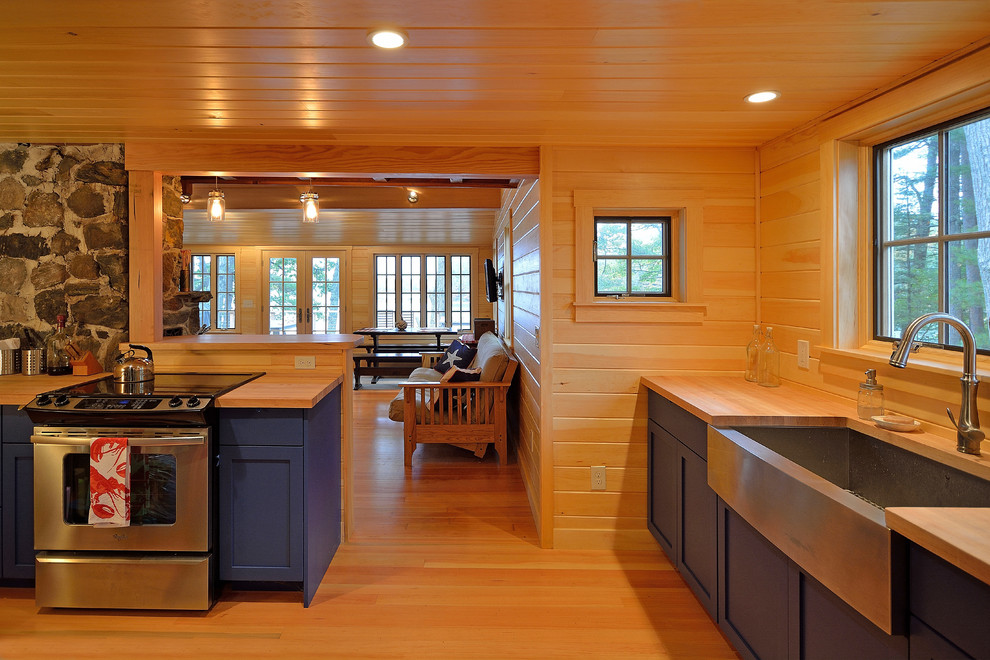 This is an example of an eclectic u-shaped open plan kitchen in Portland Maine with a farmhouse sink, blue cabinets, wood benchtops, stainless steel appliances and medium hardwood floors.