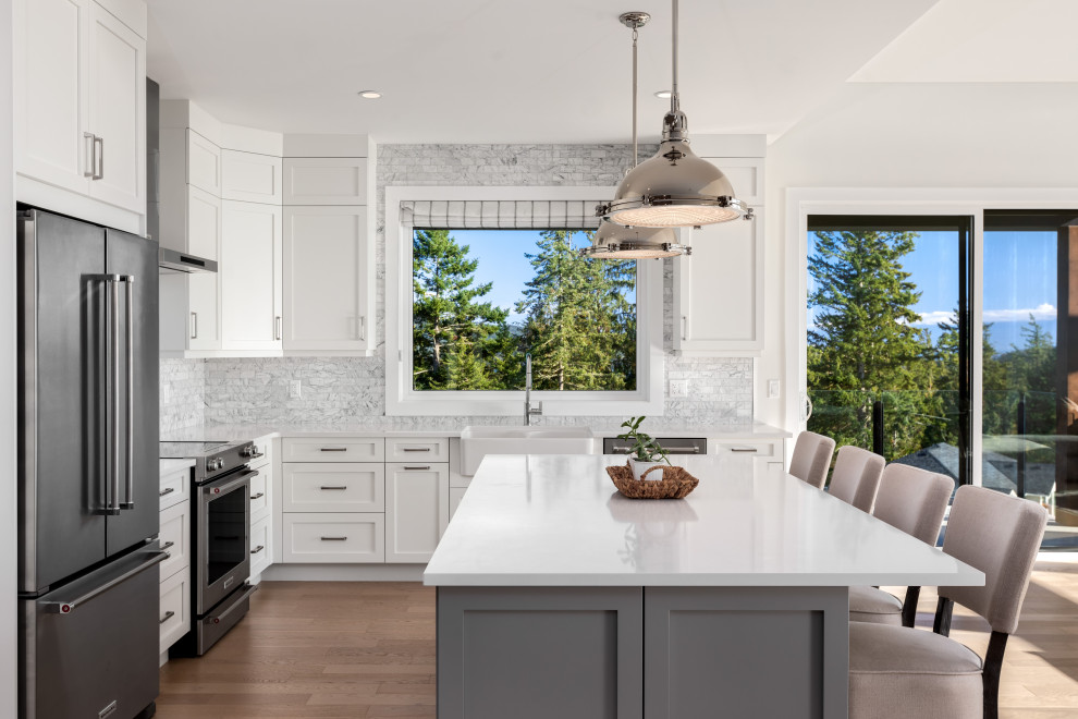 This is an example of a transitional l-shaped kitchen in Other with a farmhouse sink, shaker cabinets, white cabinets, grey splashback, stainless steel appliances, light hardwood floors, with island, beige floor and white benchtop.