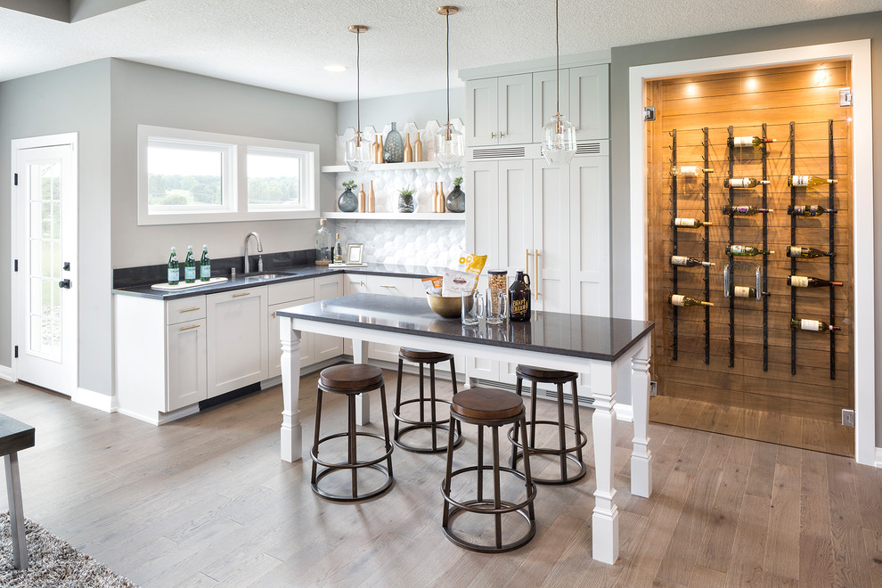 Inspiration for a transitional l-shaped seated home bar in Minneapolis with an undermount sink, shaker cabinets, white cabinets, white splashback, light hardwood floors and black benchtop.