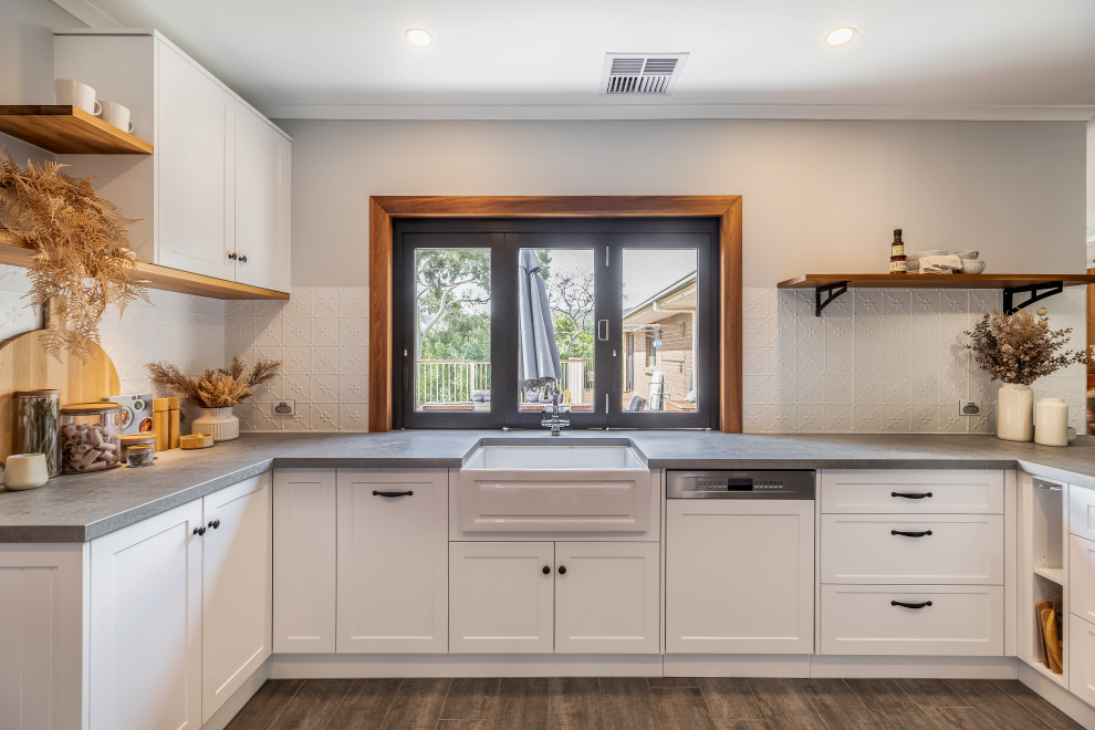 This is an example of a large rural u-shaped kitchen pantry in Adelaide with a belfast sink, shaker cabinets, white cabinets, engineered stone countertops, white splashback, metal splashback, black appliances, ceramic flooring, a breakfast bar, grey floors and grey worktops.