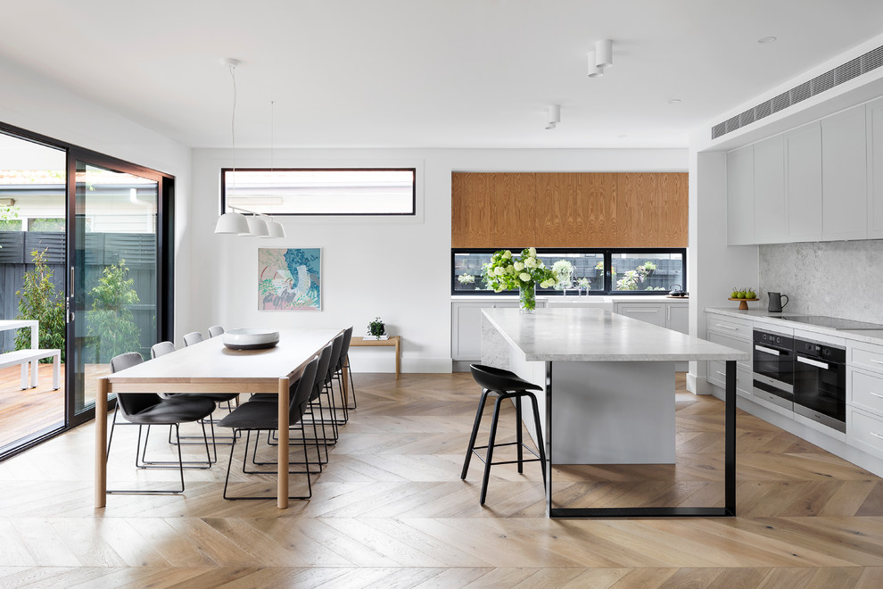 This is an example of a large beach style eat-in kitchen in Melbourne with grey cabinets, quartz benchtops, grey splashback, ceramic splashback, light hardwood floors, with island, grey benchtop, a farmhouse sink, shaker cabinets, black appliances and beige floor.