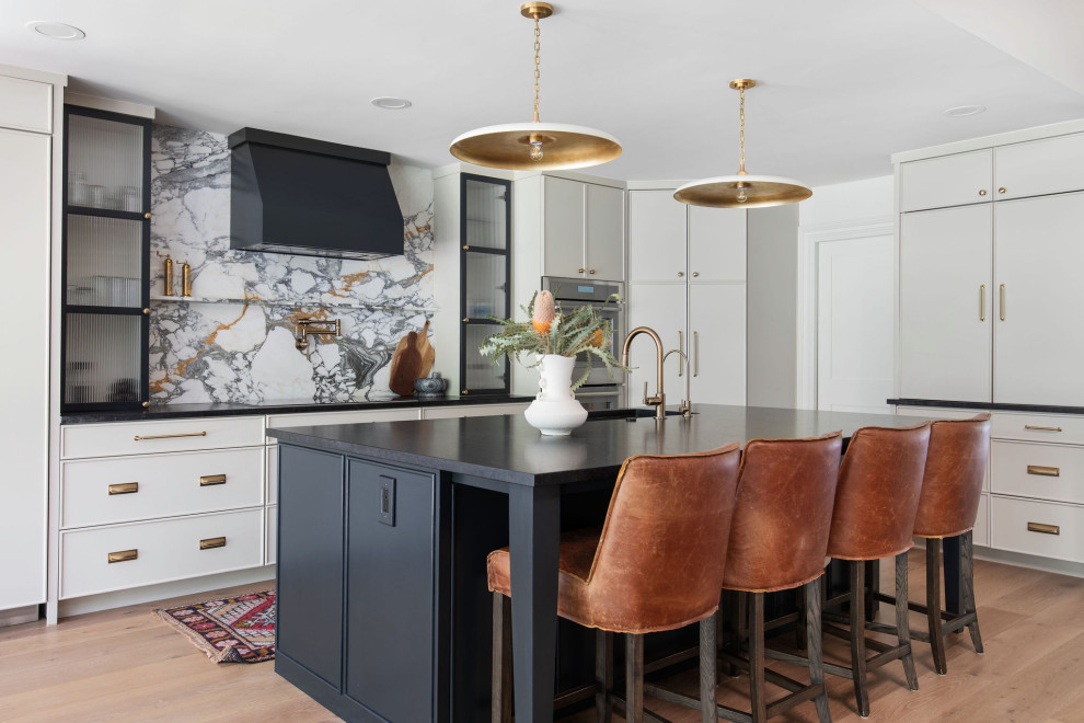 Example of a transitional l-shaped medium tone wood floor and brown floor kitchen design in Austin with shaker cabinets, white cabinets, gray backsplash, stone slab backsplash, stainless steel appliances, an island and black countertops