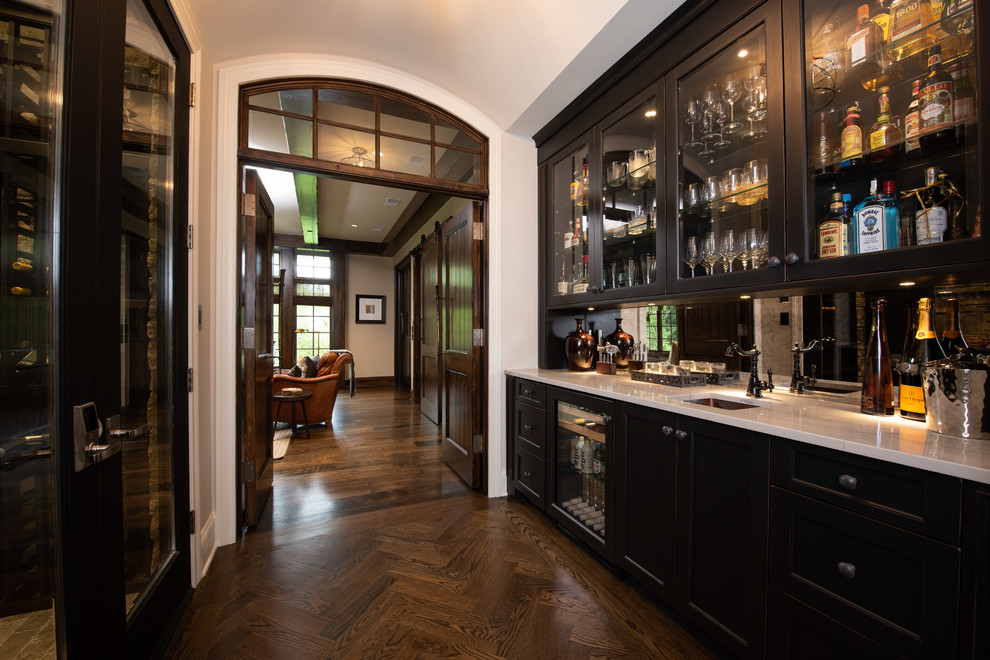 Photo of a large traditional galley wet bar in Chicago with an undermount sink, glass-front cabinets, dark wood cabinets, quartzite benchtops, mirror splashback, medium hardwood floors, brown floor and white benchtop.