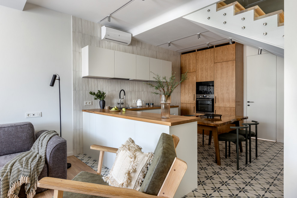 This is an example of a scandinavian u-shaped open plan kitchen with flat-panel cabinets, white cabinets, wood benchtops, grey splashback, a peninsula, multi-coloured floor, brown benchtop and a drop-in sink.
