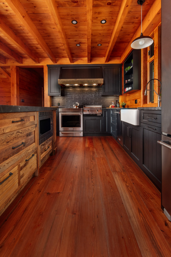 Mid-sized mountain style l-shaped medium tone wood floor and brown floor eat-in kitchen photo with a farmhouse sink, shaker cabinets, black cabinets, quartz countertops, black backsplash, stainless steel appliances, an island and black countertops