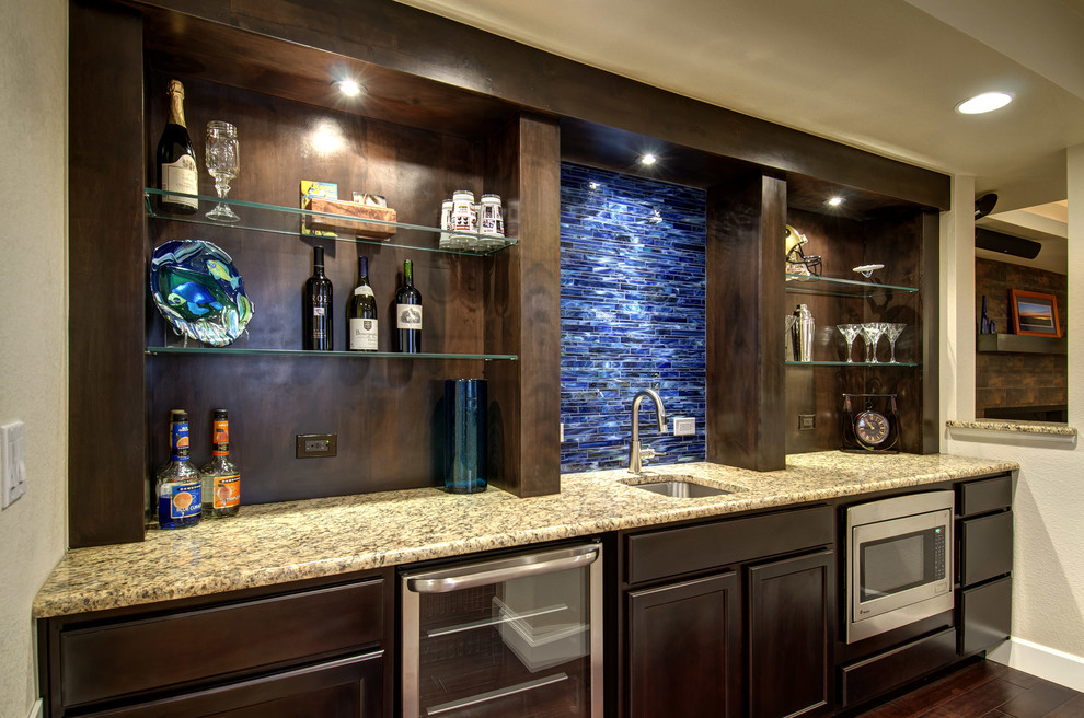 This is an example of a large transitional u-shaped seated home bar in Denver with an undermount sink, raised-panel cabinets, dark wood cabinets, granite benchtops, blue splashback, glass tile splashback, dark hardwood floors, brown floor and beige benchtop.