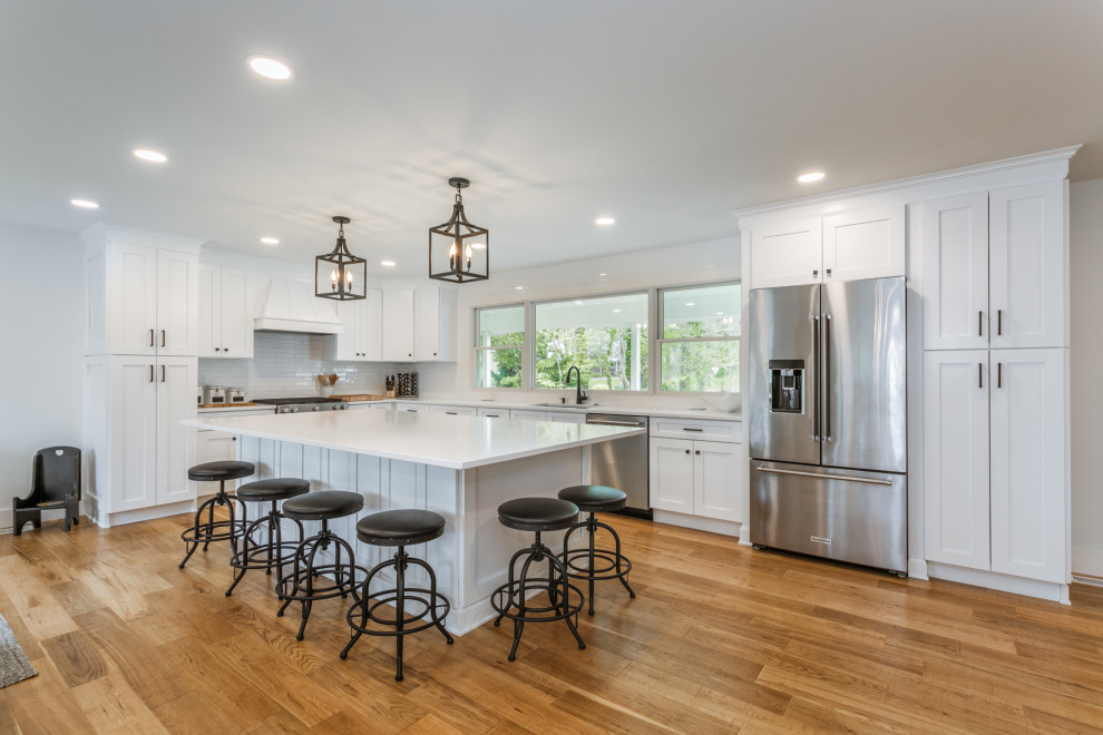 Photo of a large country l-shaped open plan kitchen in Burlington with an integrated sink, shaker cabinets, white cabinets, quartz benchtops, white splashback, ceramic splashback, stainless steel appliances, light hardwood floors, with island, brown floor and white benchtop.
