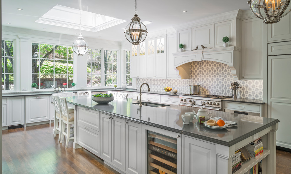 This is an example of a mid-sized traditional u-shaped open plan kitchen in Boston with raised-panel cabinets, white cabinets, stainless steel appliances, medium hardwood floors, with island, a farmhouse sink, solid surface benchtops, grey splashback, cement tile splashback and brown floor.