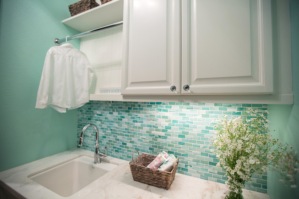 This is an example of a mid-sized beach style galley dedicated laundry room in Los Angeles with an undermount sink, raised-panel cabinets, white cabinets, marble benchtops, blue walls, porcelain floors, a stacked washer and dryer and beige floor.