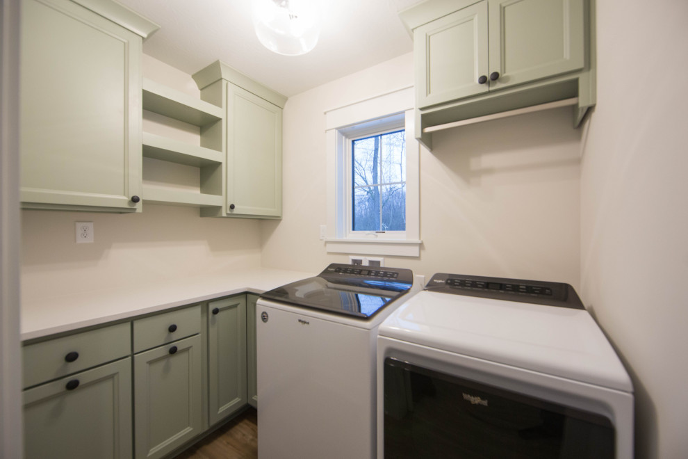 This is an example of a mid-sized contemporary l-shaped dedicated laundry room in Indianapolis with recessed-panel cabinets, green cabinets, white walls, medium hardwood floors, a side-by-side washer and dryer, brown floor and white benchtop.