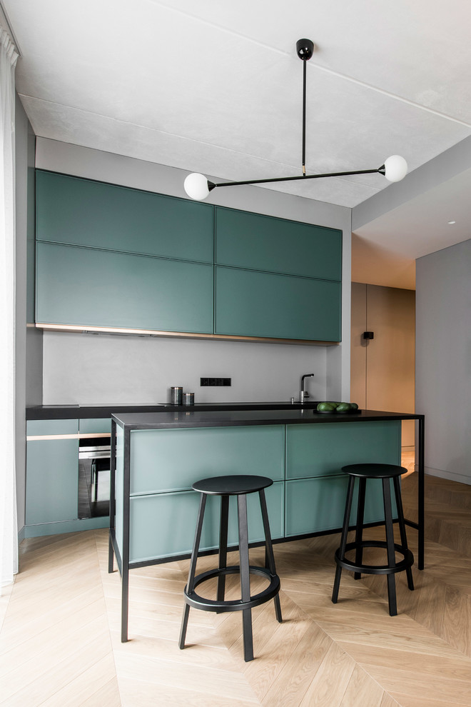 This is an example of a contemporary galley kitchen in New York with flat-panel cabinets, green cabinets, grey splashback, light hardwood floors, with island, beige floor and black benchtop.