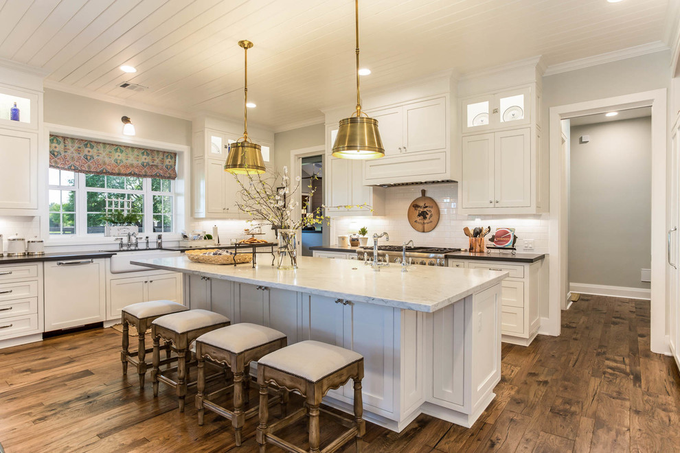 Design ideas for a country kitchen in Houston with a farmhouse sink, white cabinets, white splashback, medium hardwood floors, with island, brown floor, shaker cabinets, subway tile splashback and stainless steel appliances.