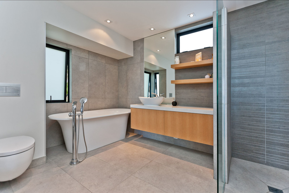 Mid-sized contemporary 3/4 bathroom in Auckland with beaded inset cabinets, light wood cabinets, a freestanding tub, a corner shower, a wall-mount toilet, gray tile, ceramic tile, grey walls, porcelain floors, a pedestal sink and laminate benchtops.