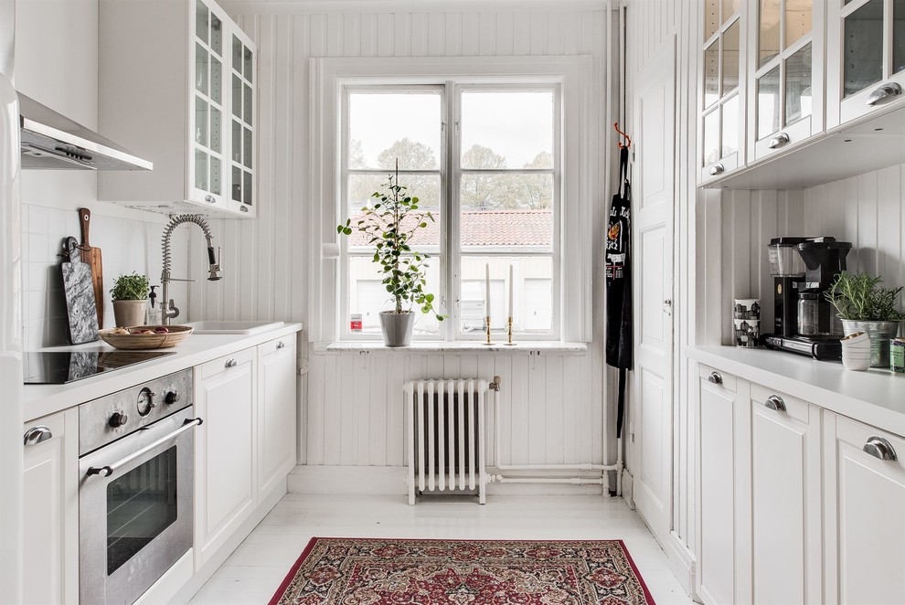 This is an example of a mid-sized country galley kitchen in Gothenburg with glass-front cabinets, white splashback, stainless steel appliances, no island, white floor and white cabinets.