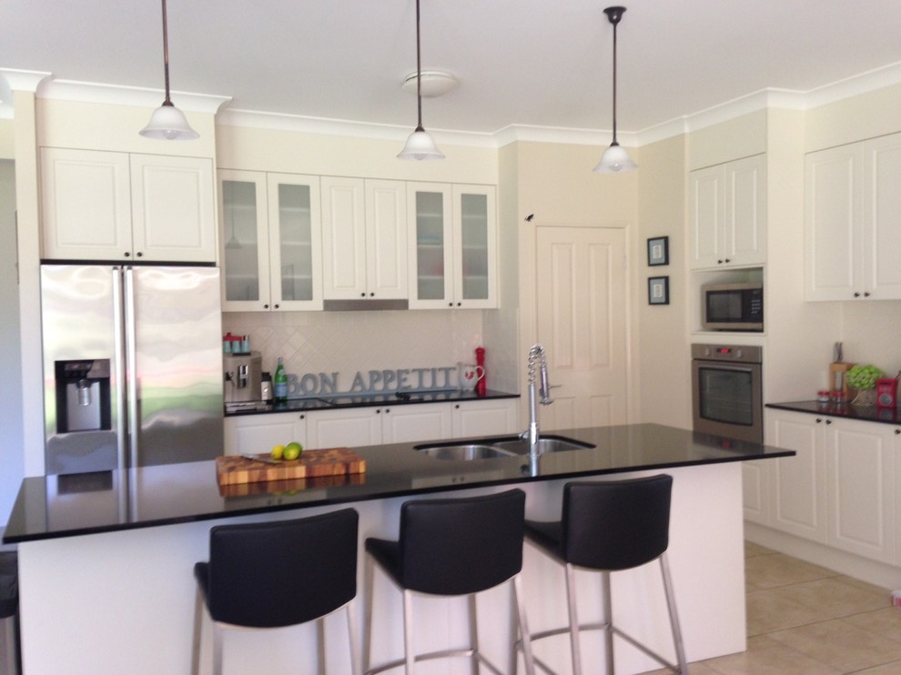 This is an example of a large transitional l-shaped eat-in kitchen in Brisbane with an undermount sink, white cabinets, quartz benchtops, white splashback, ceramic splashback, stainless steel appliances, porcelain floors and with island.
