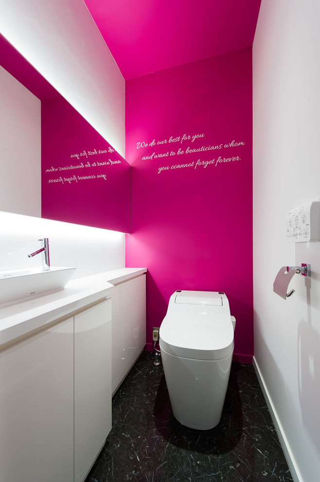 Inspiration for a mid-sized contemporary powder room in Yokohama with furniture-like cabinets, white cabinets, a one-piece toilet, pink walls, porcelain floors, a vessel sink, laminate benchtops, black floor and white benchtops.