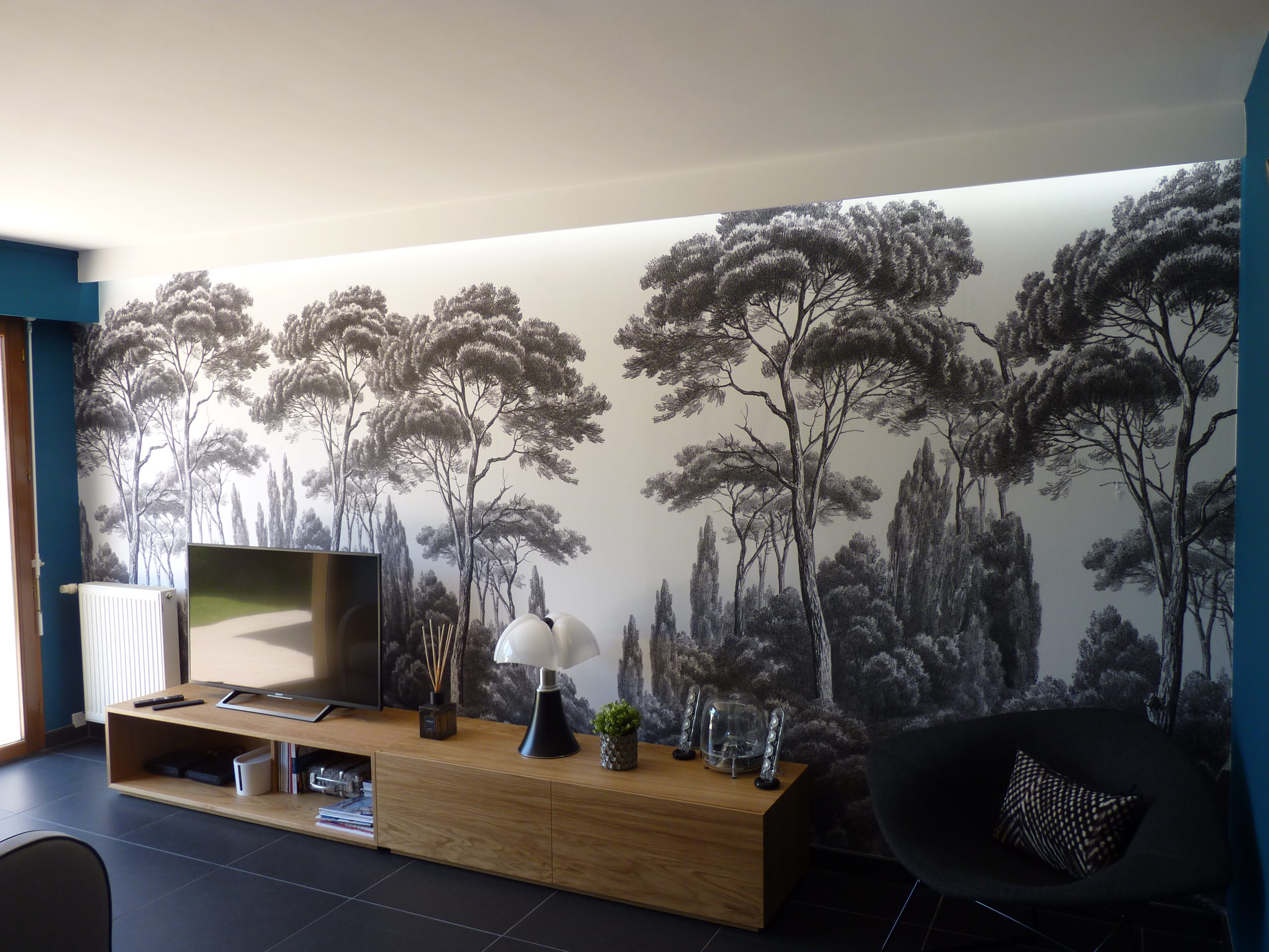 Décoration d'un salon, avec un papier peint panoramique qui habille le mur complet, mis en valeur par une lumière LED rasante indirecte. Les meubles bas permettent de ne pas trop cacher le papier pein