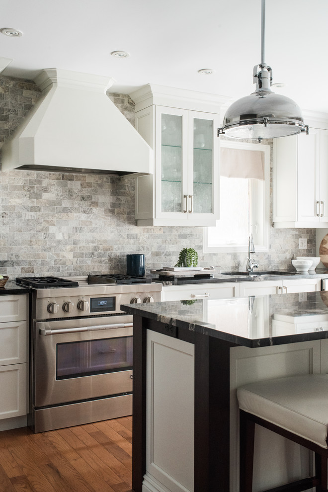 Inspiration for a transitional medium tone wood floor and brown floor kitchen remodel in Toronto with an undermount sink, recessed-panel cabinets, white cabinets, granite countertops, gray backsplash, travertine backsplash, stainless steel appliances and an island
