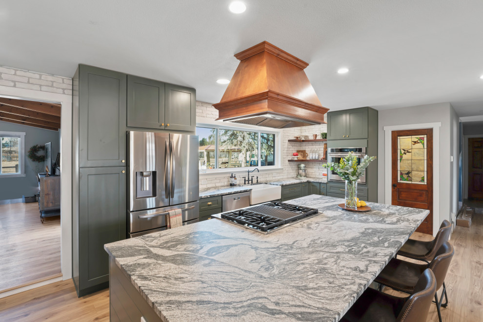 Lantlig inredning av ett stort grå grått kök, med en rustik diskho, skåp i shakerstil, gröna skåp, granitbänkskiva, vitt stänkskydd, stänkskydd i tegel, rostfria vitvaror, vinylgolv, en köksö och beiget golv