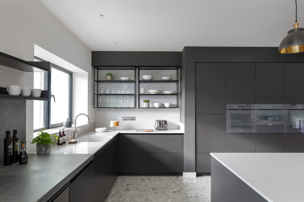 This is an example of an expansive contemporary l-shaped open plan kitchen in Sussex with flat-panel cabinets, black cabinets, with island, a single-bowl sink, panelled appliances, grey floor and white benchtop.